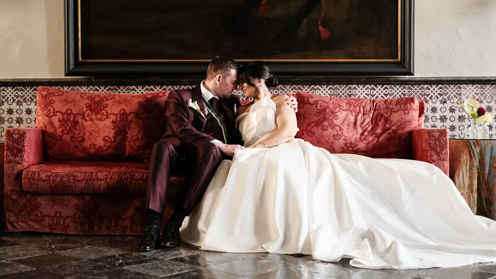 Brautpaar sitzt eng beieinander auf einem roten Sofa. Die Braut trägt ein weißes Hochzeitskleid, der Bräutigam einen dunkelroten Anzug.
