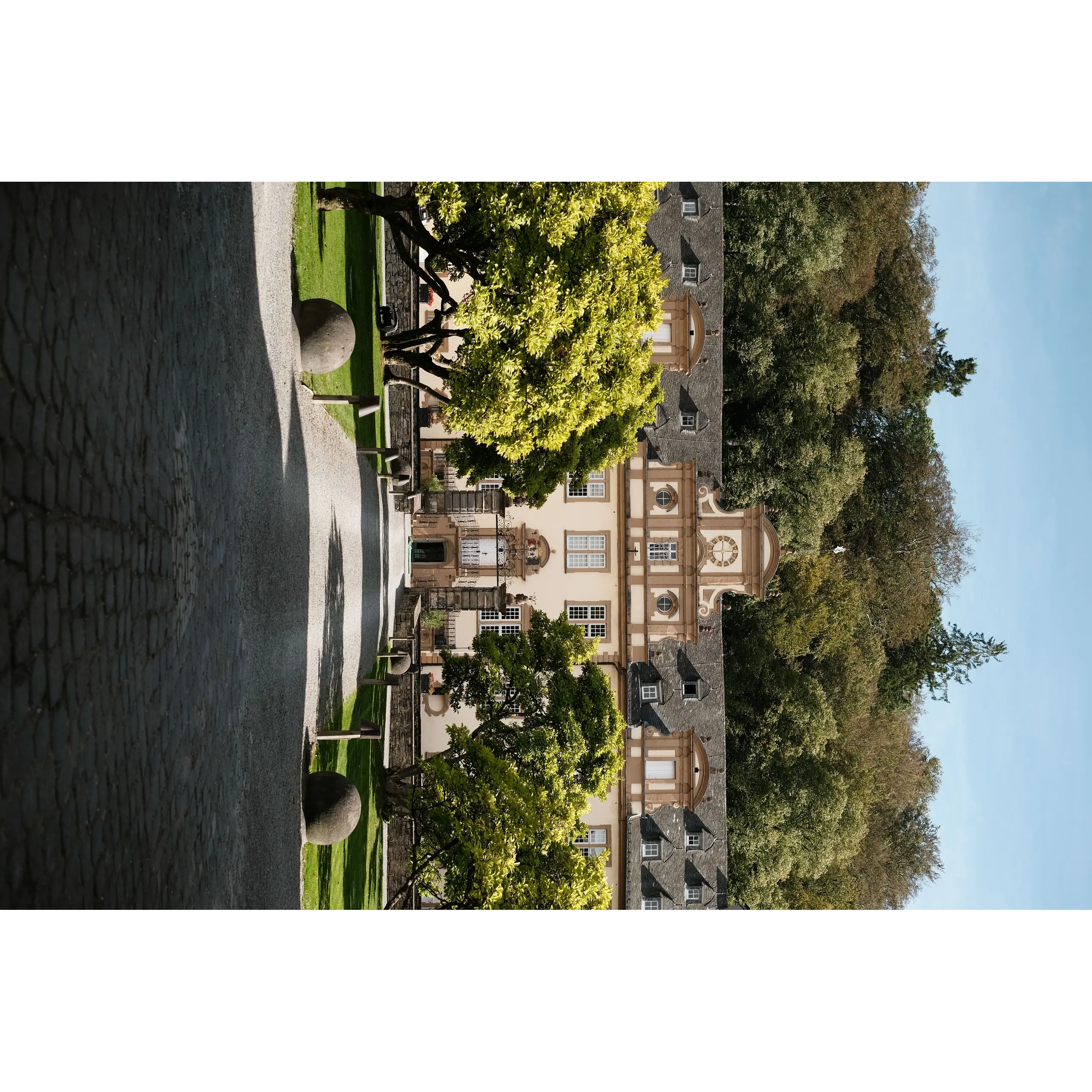 Historisches Schloss mit heller Fassade, geschwungenen Giebeln und einer von Bäumen gesäumten Zufahrt.