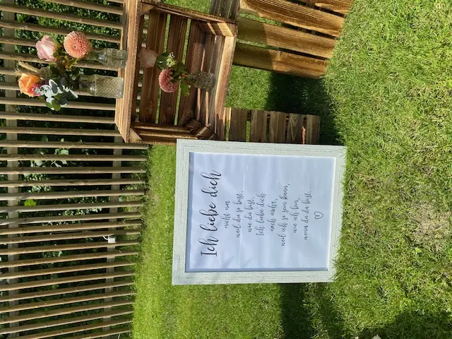 Hochzeitsdekoration im Garten mit Holzkisten, kleinen Glasvasen, rosafarbenen und orangefarbenen Blumen sowie einem gerahmten Liebesspruch.
