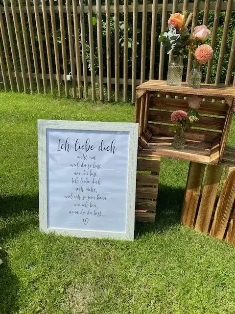 Hochzeitsdekoration im Garten mit Holzkisten, kleinen Glasvasen, rosafarbenen und orangefarbenen Blumen sowie einem gerahmten Liebesspruch.